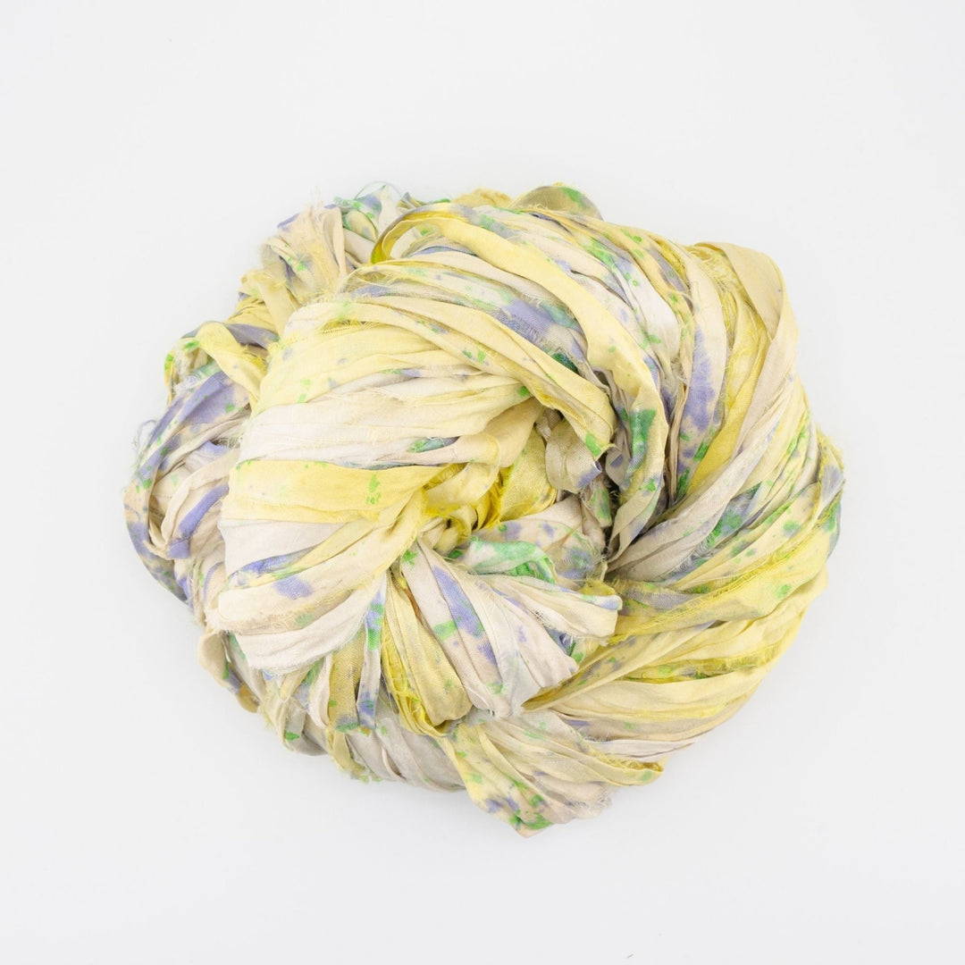 Close-up image of a skein of small batch sari silk ribbon yarn, featuring a mix of earthy tones including yellow and green. The ribbon displays a unique, textured appearance characteristic of upcycled sari silk. Ideal for use in knitting, crocheting, weaving, and mixed media projects.