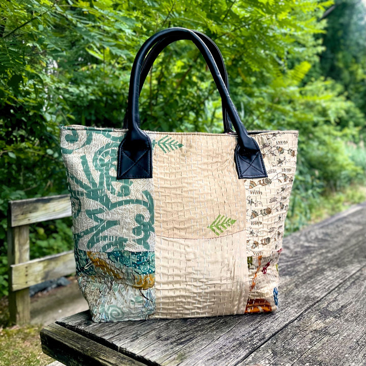 A boho cotton patchwork purse sitting on a wooden table outside. The bag features kantha stitching and sturdy leather handles.