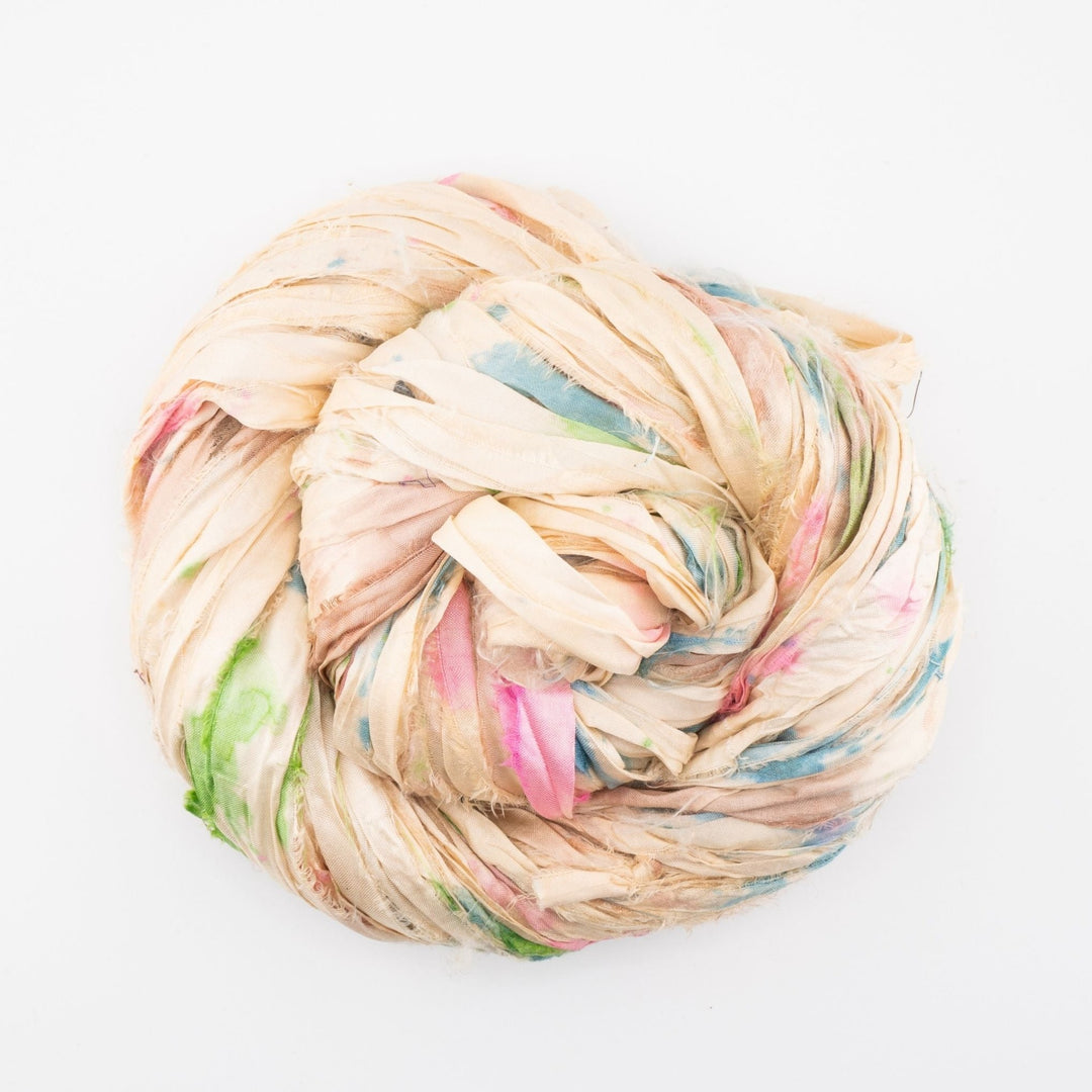 Close-up image of a skein of small batch sari silk ribbon yarn, featuring soft pastel colors including cream, pink, and blue. The ribbon has a unique, textured look that is characteristic of upcycled sari silk. Ideal for various crafting projects such as knitting, crocheting, weaving, and mixed media.