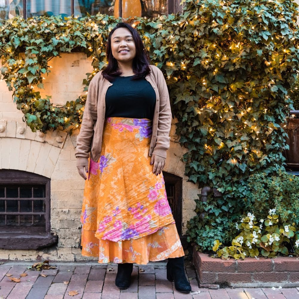 Woman in an orange skirt and tan cardigan standing in front of covered wall with lights