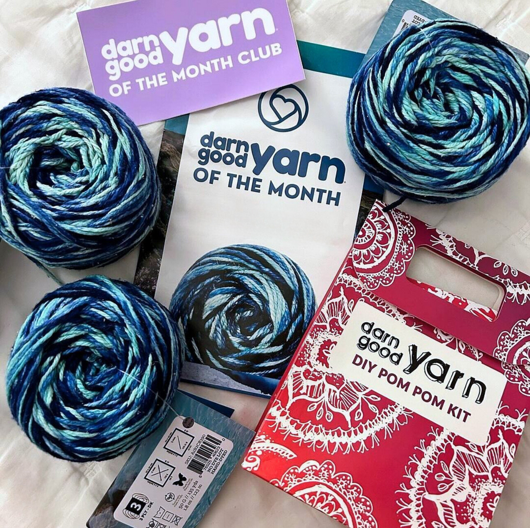 A word Yarn of the Month Club box with blue skeins of yarn and a DIY pom pom kit