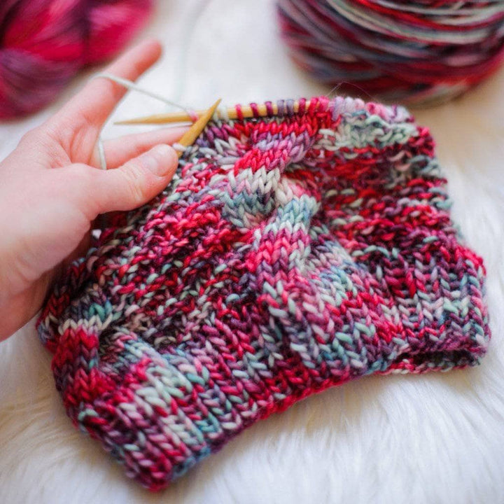 Partially knit Ash Cave Beanie in Benjamin (red and gray) on circular knitting needles with a yarn cake a white fur background