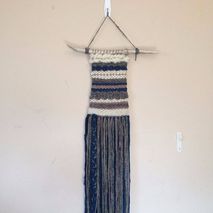 Woven tapestry in gray, navy, and white hanging from wooden stick on a white wall