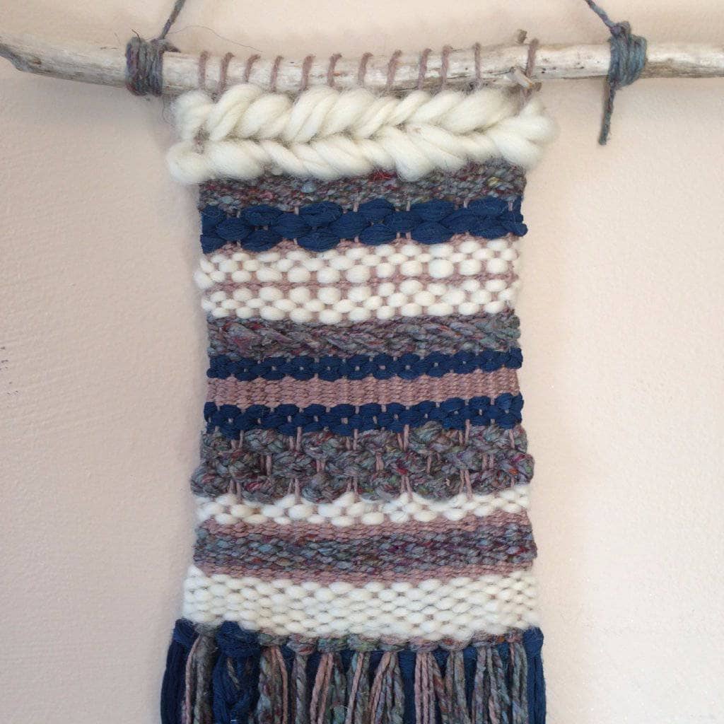 Woven tapestry in gray, navy, and white hanging from wooden stick on a white wall