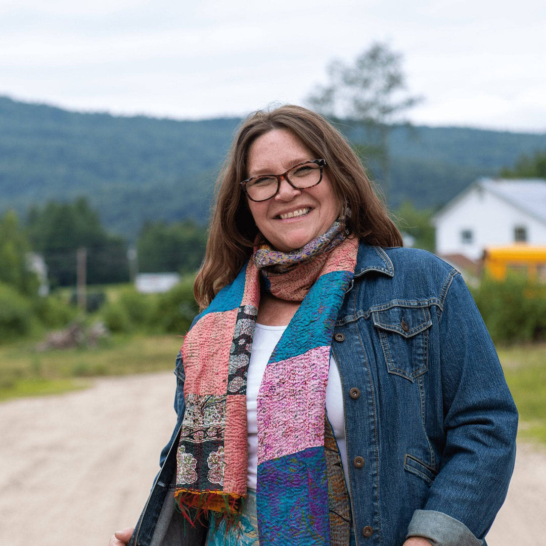 Model wearing one of a kind kantha scarf (pink and blue) outside with a farm house and hill in the background.