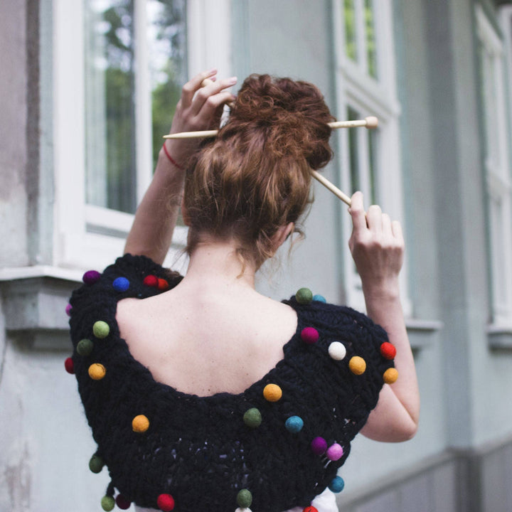 A photo of a woman from behind. She's wearing a CaPowl Cowl in the color black. She has knitting needles in her hair.