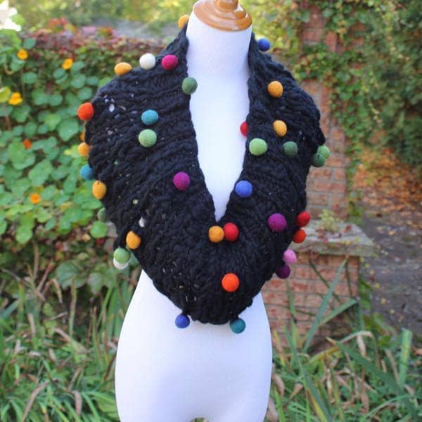 Mannequin wearing a Capowl Cowl in black with multicolored felt ball accents standing in front of greenery