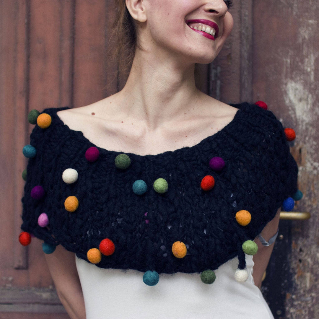 A woman standing against a wooden door wearing a CaPowl Felt Ball cowl in black.