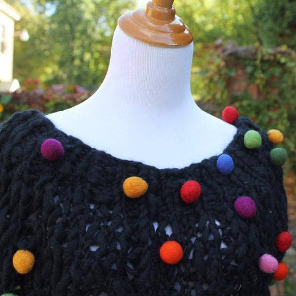 Mannequin wearing a Capowl Cowl in black with multicolored felt ball accents standing in front of greenery