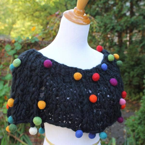 Mannequin wearing a Capowl Cowl in black with multicolored felt ball accents standing in front of greenery