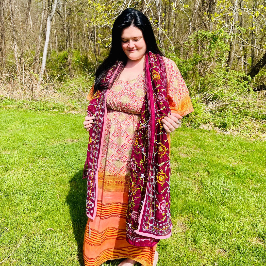 Woman wearing a traditional dress outdoors, holding a richly embroidered shawl.