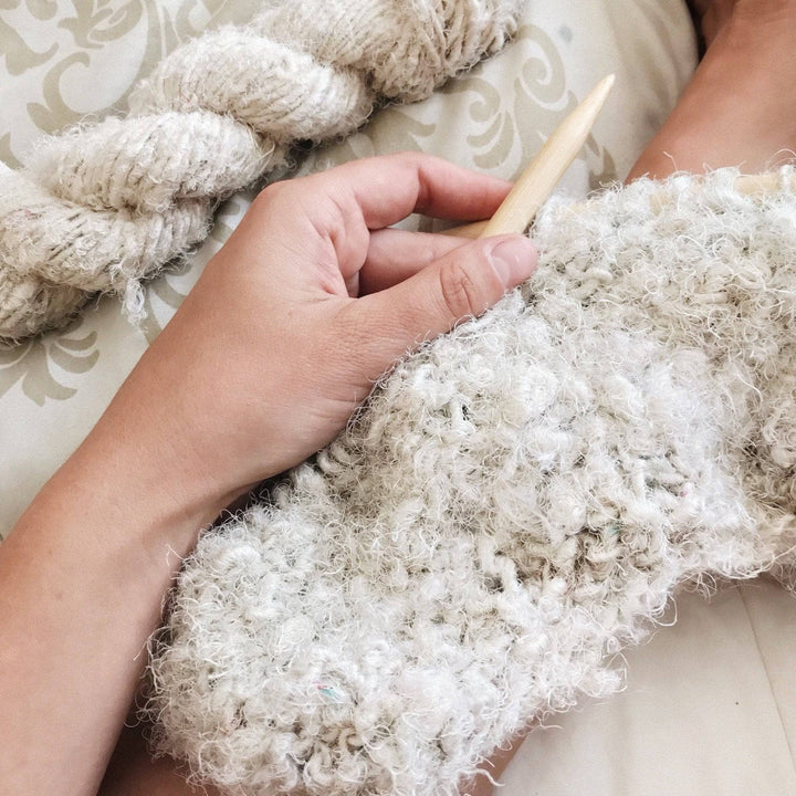 Woman's hand knitting a cream colored knit cowl on a beige surface