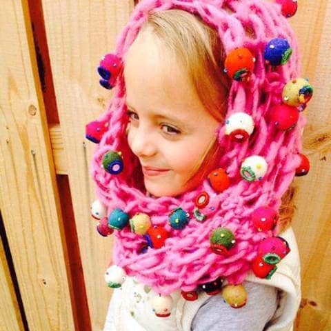 Little girl wearing a pink cowl hat with felt ball embellishments standing in front of a wooden fence