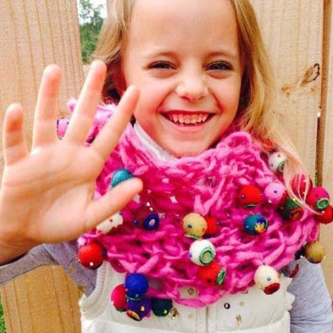 Little girl wearing a pink cowl hat with felt ball embellishments standing in front of a wooden fence