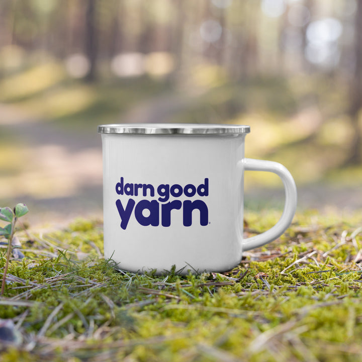 A white enamel mug with "darn good yarn" printed, set on a mossy forest floor.