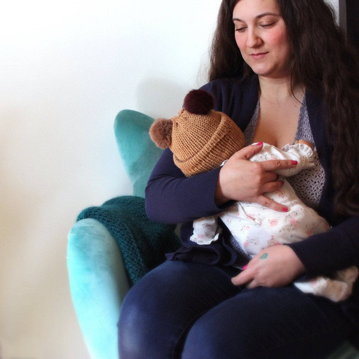 Baby wearing brown hat with two faux fur pom poms on top being held by woman in dark blue outfit upon a light blue chair in front of a blank wall.