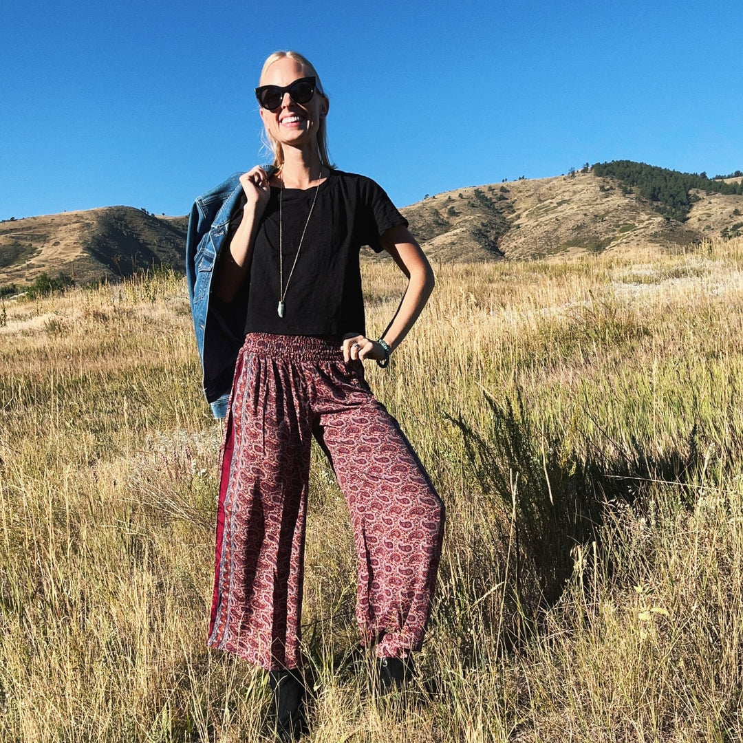 A woman smiling in a field, dressed in red printed pants and a black top.