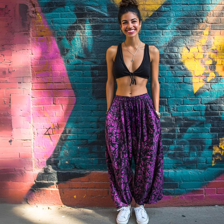 A Woman in purple patterned pants and a black top, smiling in front of painted brick wall.