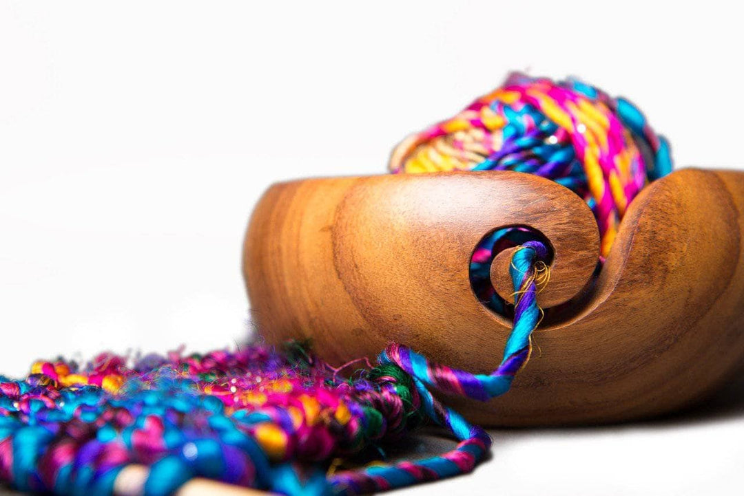 Wooden brown yarn bowl with swirl cutout for yarn ends filled with multicolored yarn and sitting on a white surface