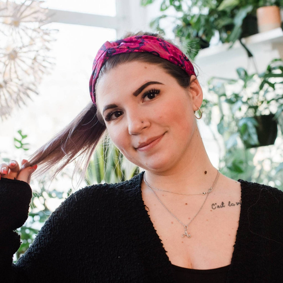 Model is wearing a pink kameela not headband in her hair while standing in front of some green plants. 