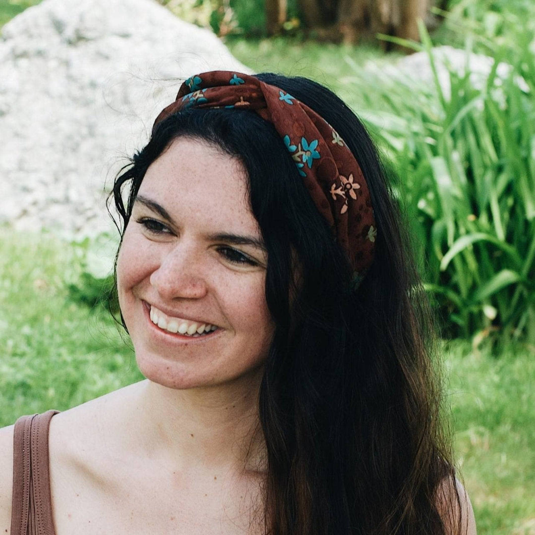 Woman is smiling sitting in a garden wearing a brown sari silk kameela knot headband with blue florals on it.