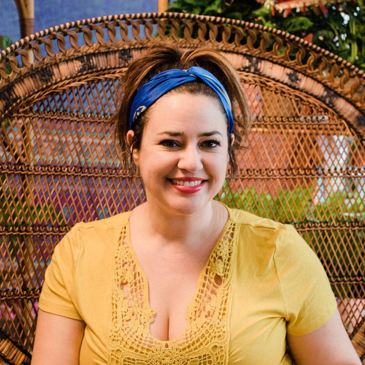 Woman is sitting in a big papasan chair with an ocean blue sari silk kameela knot headband in her hair. There's gold symbols on the headband.