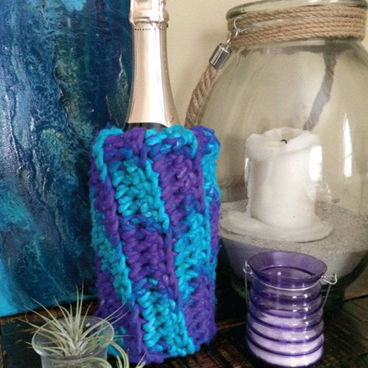 wine bottle covered in a bulky sleeve next to two candles a plant with a blue and white background