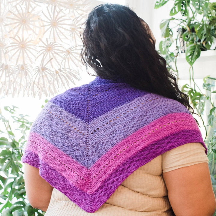 model wearing purple ombre stitch sampler shawl over their shoulders with potted greenery in the background.