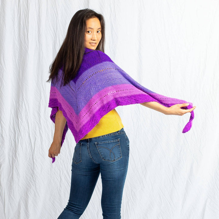 Model wearing ombre stitch sampler shawl in purple outstretched in front of a white background.