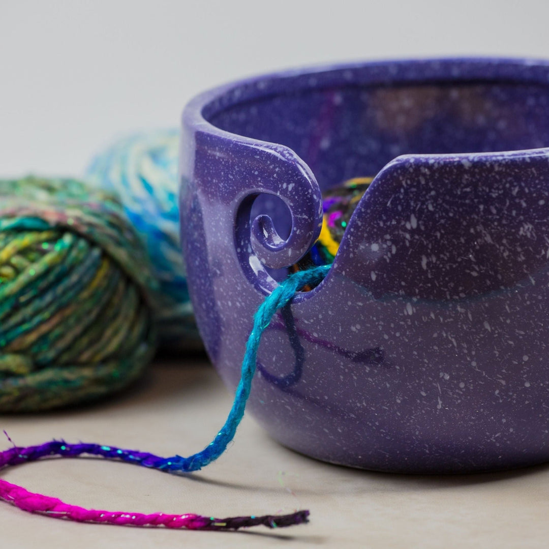 Purple Cosmic Ceramic Yarn Bowl with white speckles, holding multicolored yarn, with two balls of green and blue yarn in the background. This deep-space-inspired yarn bowl helps keep yarn clean, organized, and untangled during knitting or crochet projects.