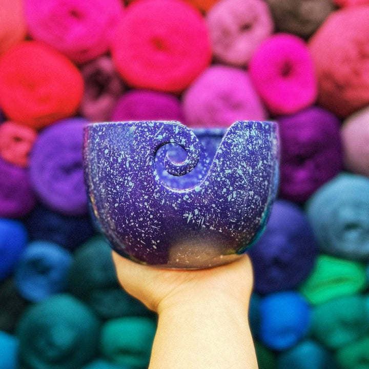 Hand holding a Purple Cosmic Ceramic Yarn Bowl with white speckles in front of a background of colorful yarn balls. This celestial-themed yarn bowl helps keep yarn clean, organized, and untangled during knitting or crochet projects.