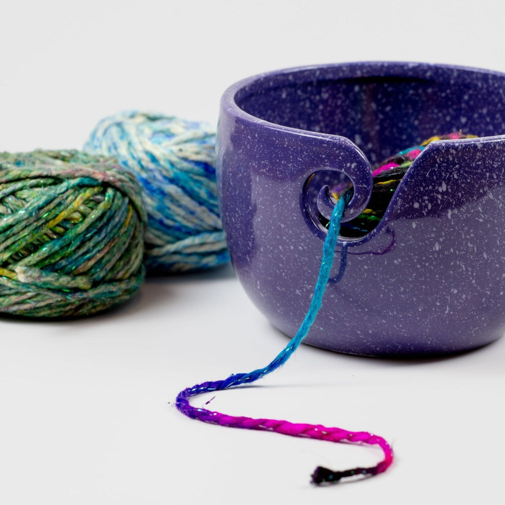 Purple Cosmic Ceramic Yarn Bowl with white speckles, holding multicolored yarn, with two balls of green and blue yarn in the background. This celestial-themed yarn bowl helps keep yarn clean, organized, and untangled during knitting or crochet projects.