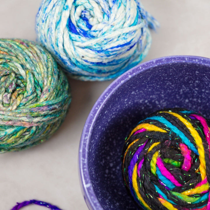 Top view of a Purple Cosmic Ceramic Yarn Bowl with white speckles, holding a ball of multicolored yarn, with two additional balls of blue and green yarn beside it. This celestial-themed yarn bowl helps keep yarn clean, organized, and untangled during knitting or crochet projects.