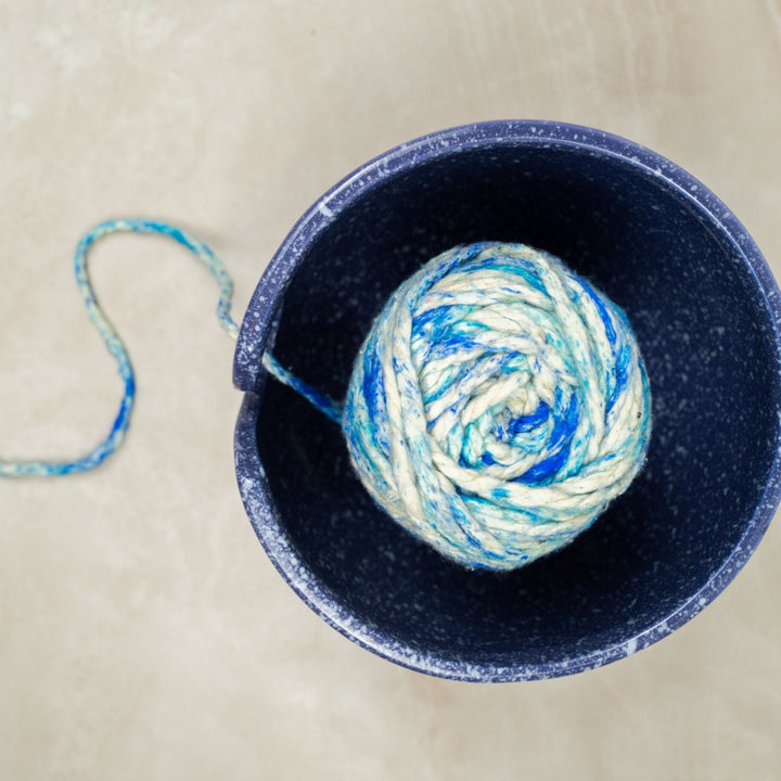 Top view of a Purple Cosmic Ceramic Yarn Bowl with white speckles, holding a ball of blue and white yarn, with the yarn threaded through the swirl on the side. This celestial-themed yarn bowl helps keep yarn clean, organized, and untangled during knitting or crochet projects.