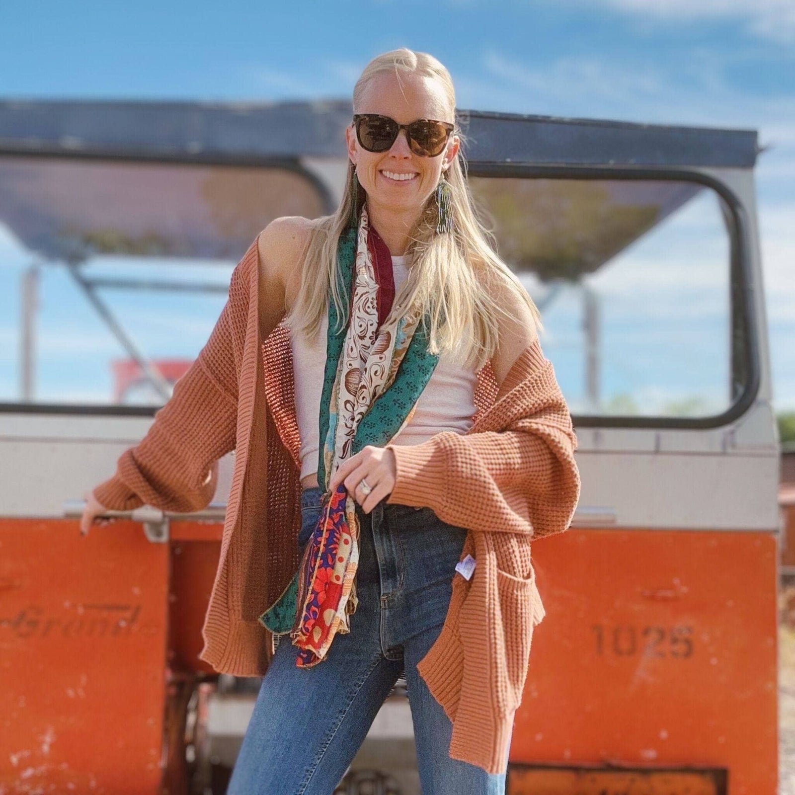 A woman posing outdoors in a cardigan and patterned scarf, standing by a vehicle.