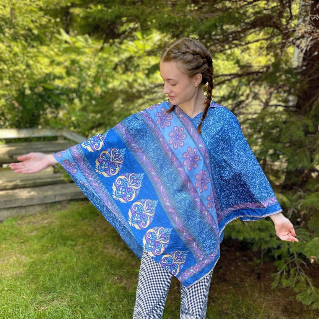 A woman in a blue patterned poncho standing outdoors near greenery.