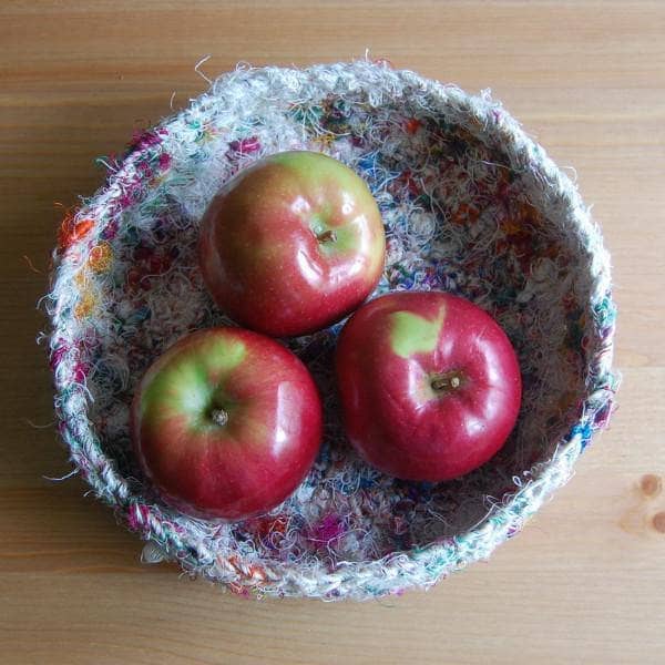 A birds eye view of a white tray made of multicolored white yarn sitting a wooden table with apples inside of the tray