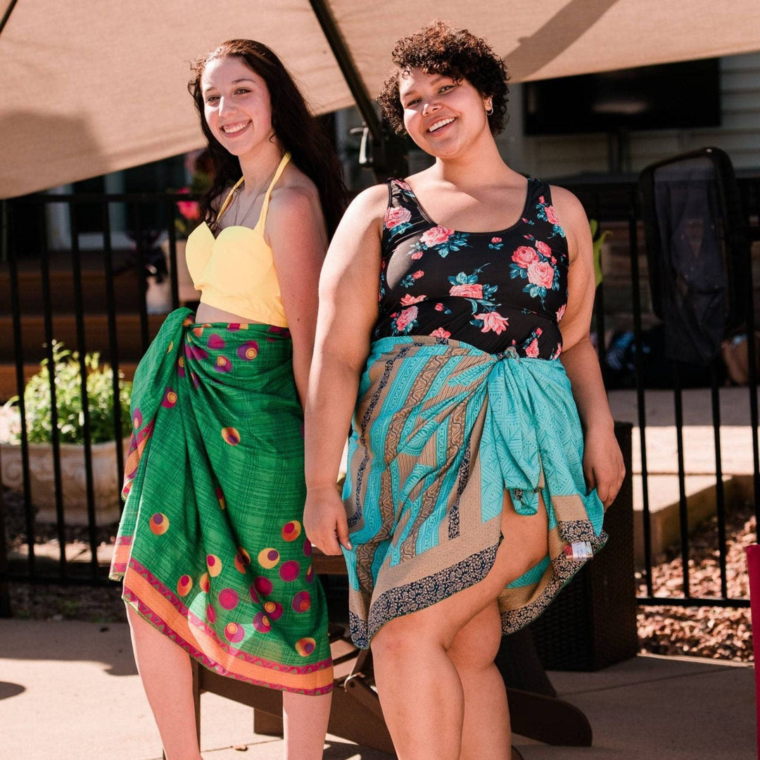 Two women standing by a pool wearing bathing suits covered up by sari silk beach sarongs. One woman is wearing a green sarong with colorful dots, the other is wearing a baby blue sarong with grey and black paisley patterns.