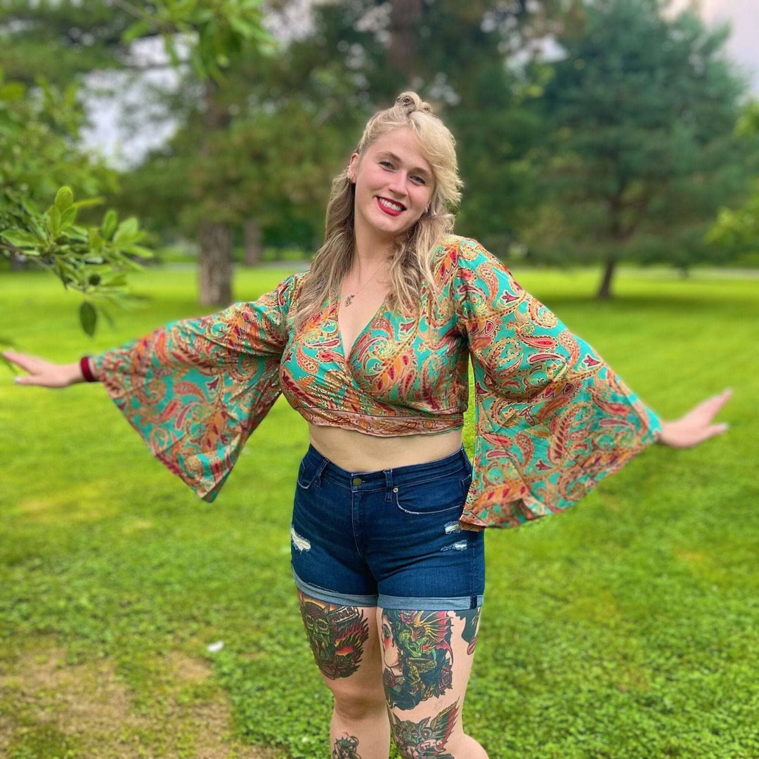 A smiling woman in a colorful top and shorts poses in a green park, showing off her tattoos