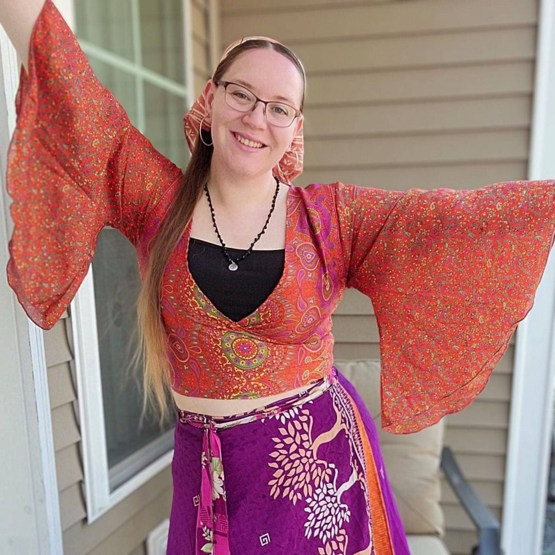 A woman smiles in front of a house wearing a colorful outfit with wide sleeves and a floral skirt.