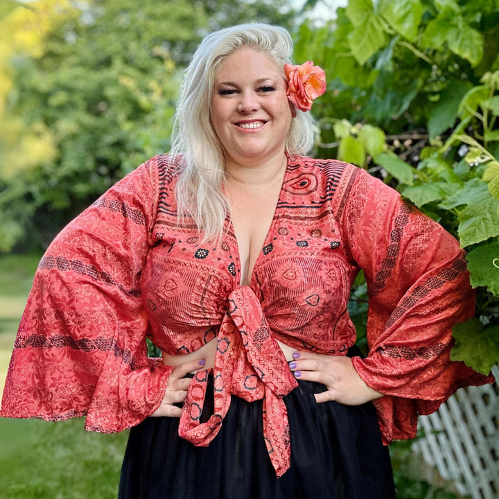 Smiling woman in a red top with flared sleeves, posing outdoors with a flower in her hair