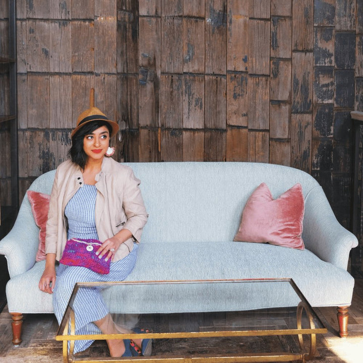 A woman holding a purple, pink and blue multicolored purse made from sari silk yarn sitting on a blue couch