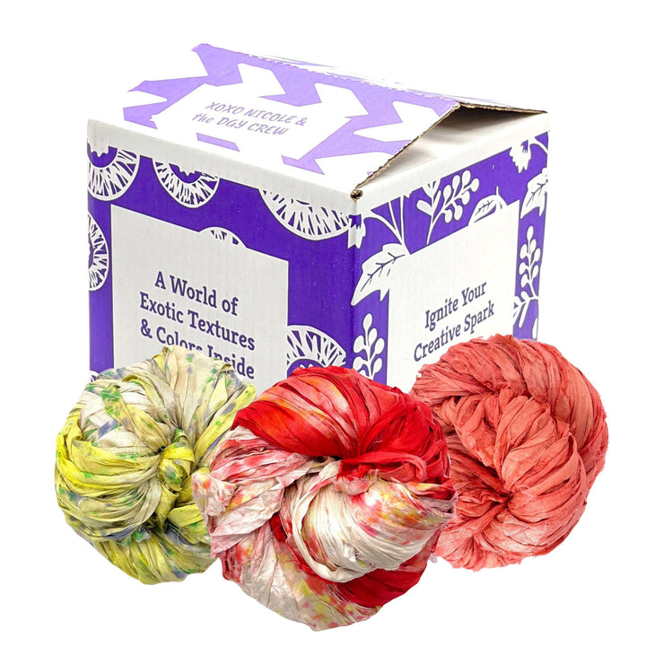 Image of the Sari Ribbon Exploration Kit, Heat Wave variant, featuring six skeins of vibrant, upcycled sari silk ribbon yarn in assorted colors. Three of the six skeins are shown in front of a decorative box with the text 'A World of Exotic Textures & Colors Inside' and 'Ignite Your Creative Spark.' The skeins display a range of rich hues: green with yellow accents, red with white and yellow accents, and peach with coral accents. Perfect for knitting, crocheting, weaving, and mixed media projects.