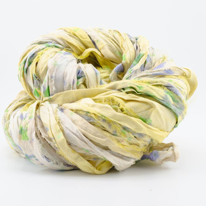 Close-up image of a skein of sari silk ribbon yarn in shades of yellow and beige, with vibrant accents of green and purple. The ribbon displays a unique, textured appearance, showcasing the varied and colorful hues characteristic of upcycled sari silk. Ideal for use in knitting, crocheting, weaving, and mixed media projects.