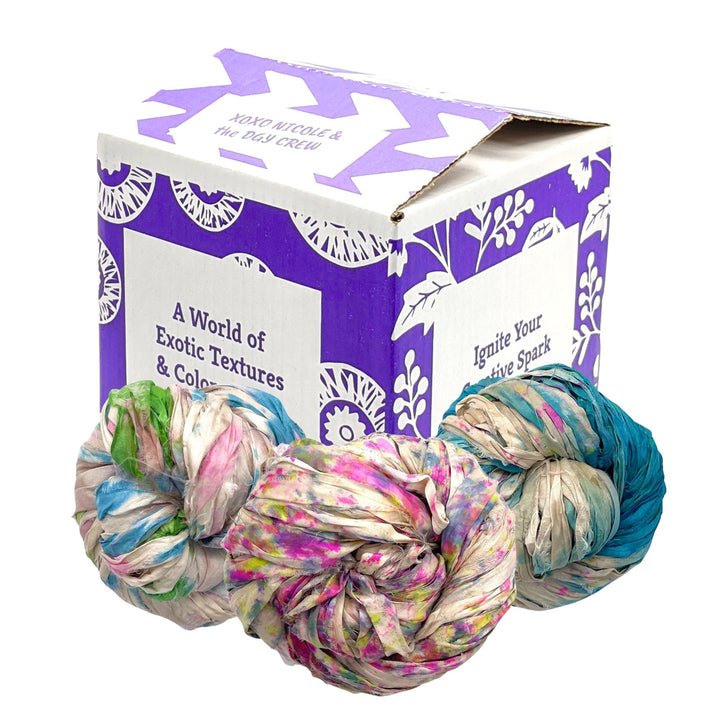 Image of the Sari Ribbon Exploration Kit, Ocean Spray variant, featuring six skeins of vibrant, upcycled sari silk ribbon yarn in assorted colors. Three of the six skeins are shown in front of a decorative box with the text 'A World of Exotic Textures & Colors Inside' and 'Ignite Your Creative Spark.' The skeins display a range of rich hues: green with pink and white accents, pink with white and green accents, and blue with beige and white accents.