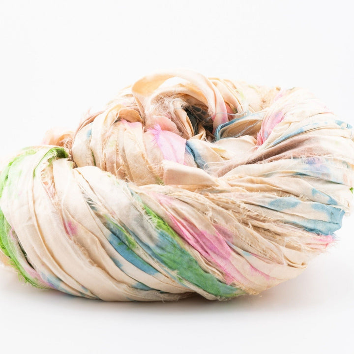 Close-up image of a skein of sari silk ribbon yarn in shades of beige, with vibrant accents of green, pink, and blue. The ribbon displays a unique, textured appearance, showcasing the varied and colorful hues characteristic of upcycled sari silk. Ideal for use in knitting, crocheting, weaving, and mixed media projects.