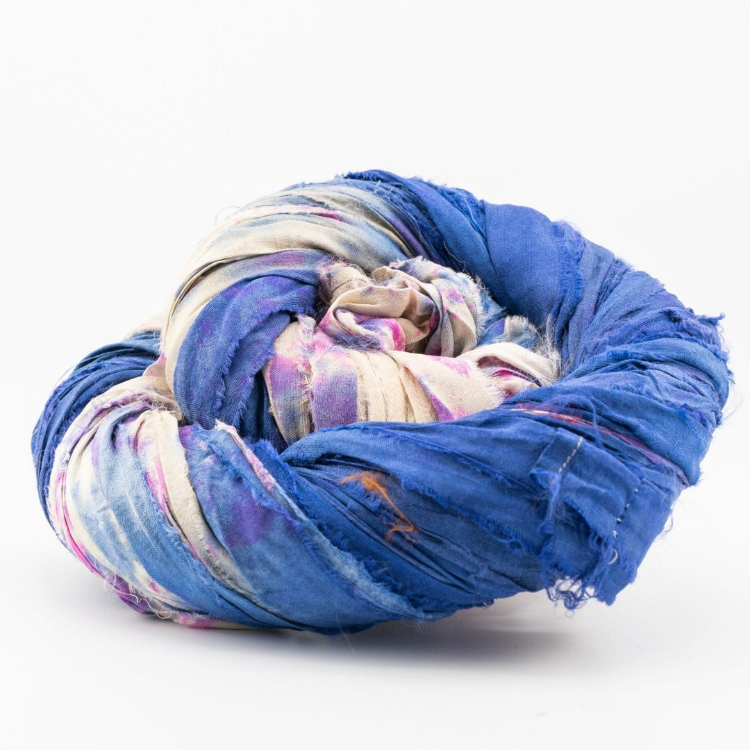 Close-up image of a skein of sari silk ribbon yarn in shades of deep blue, with vibrant accents of beige, pink, and purple. The ribbon displays a unique, textured appearance, showcasing the rich and varied hues characteristic of upcycled sari silk. Ideal for use in knitting, crocheting, weaving, and mixed media projects.