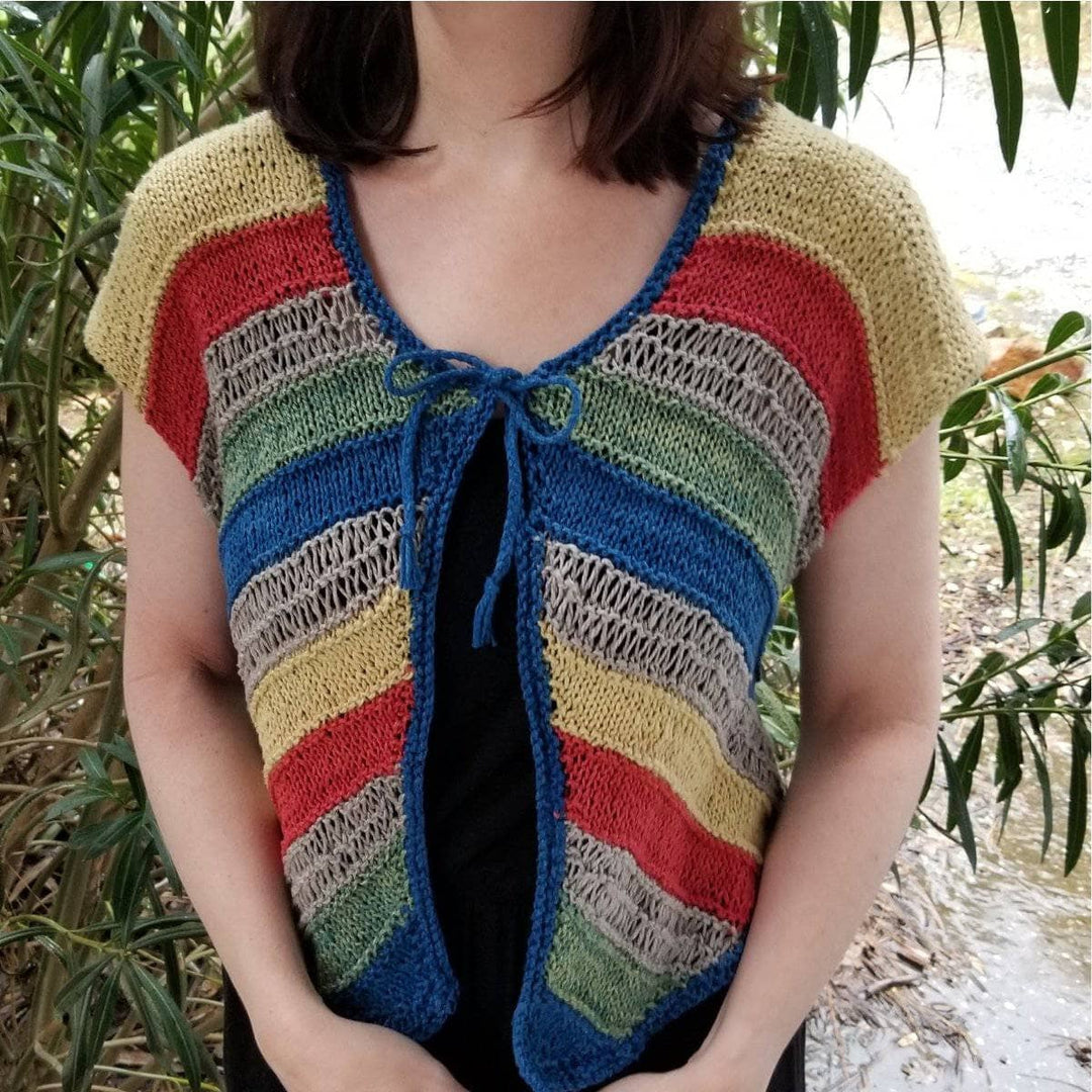 A front facing view of a women wearing a yellow, red, tan, green and blue stripped cardigan with trees in the background