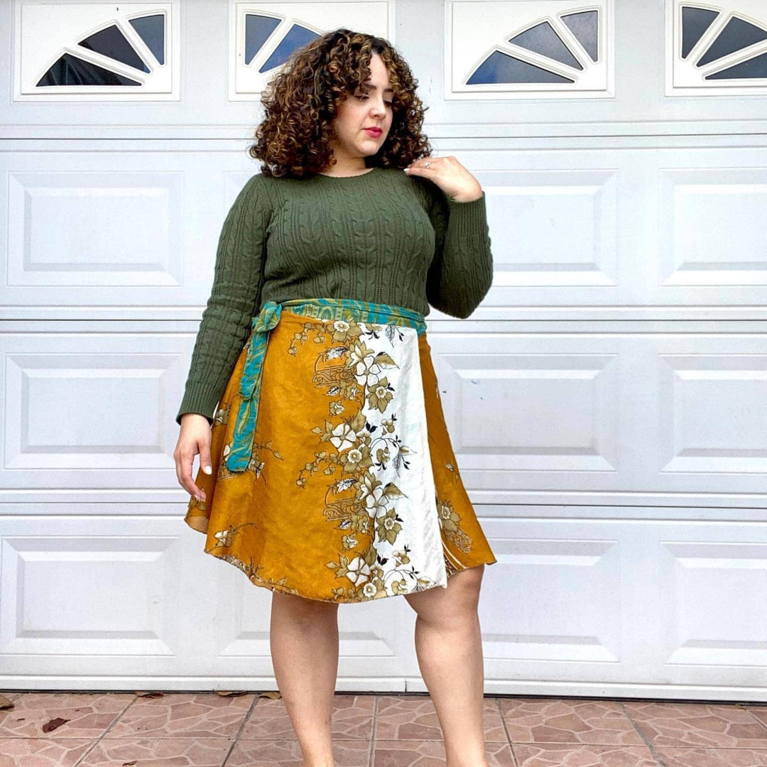 Woman with curly hair in a green sweater and floral wrap skirt stands in front of a garage door.