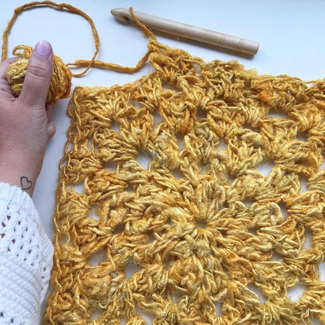 Partially made Sunny Days Tank Top with woman's hand, yarn ball, and crochet hook on a white background